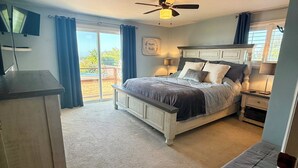 A bedroom with a bed, a desk, a chair, a ceiling fan, and a view of the outdoors.