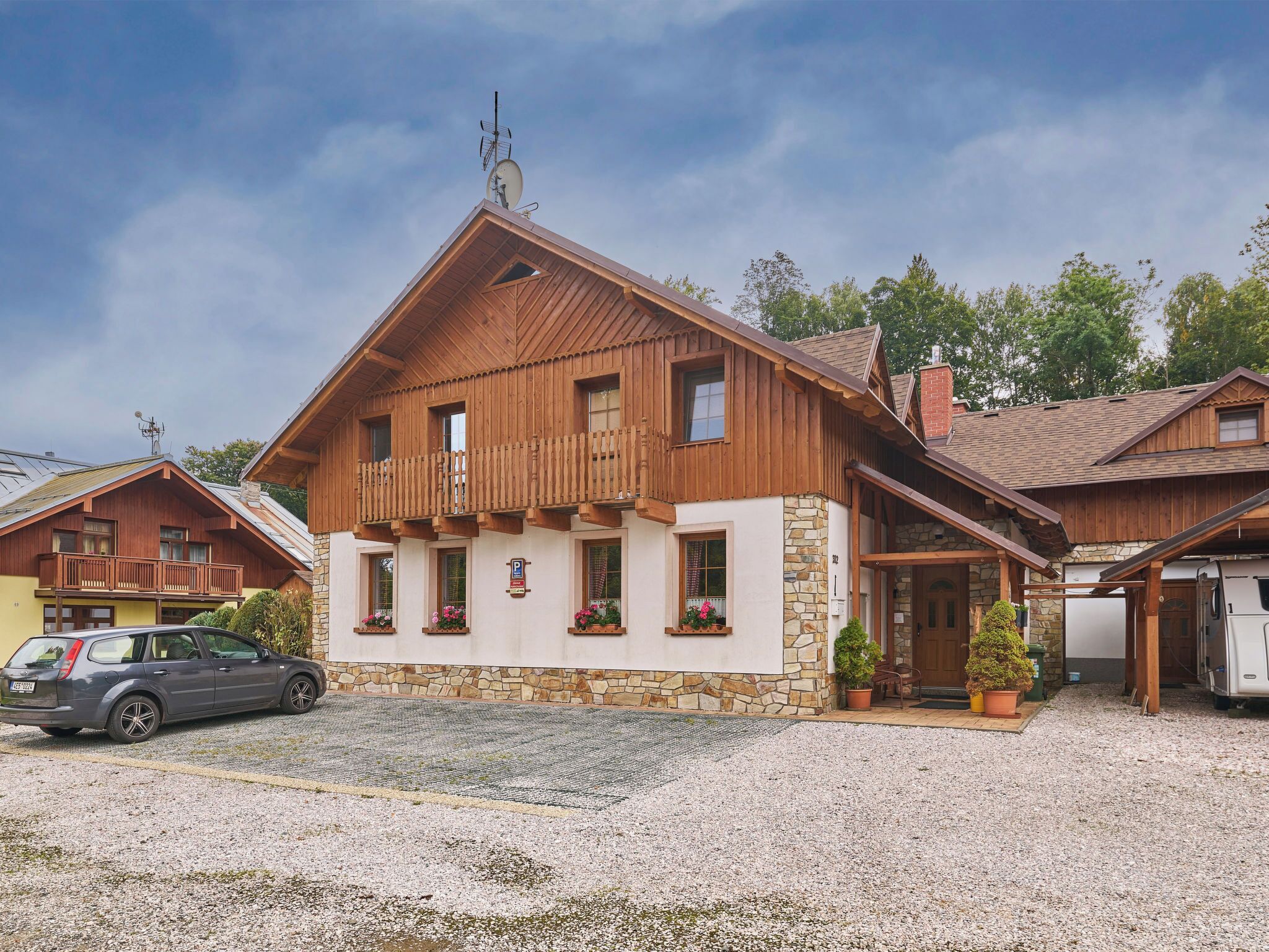 A two-story wooden house with a stone facade, a balcony, and a gravel driveway.