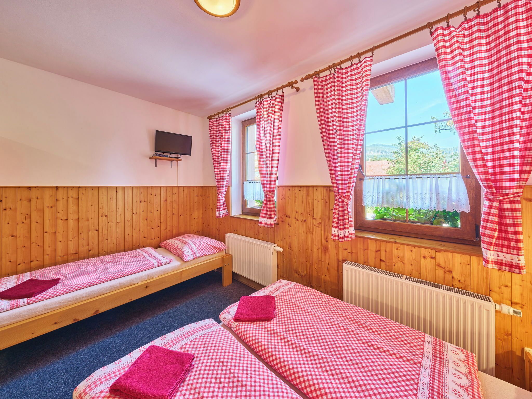 A room with three wooden bunk beds, a TV mounted on the wall, and a window with red and white checkered curtains.