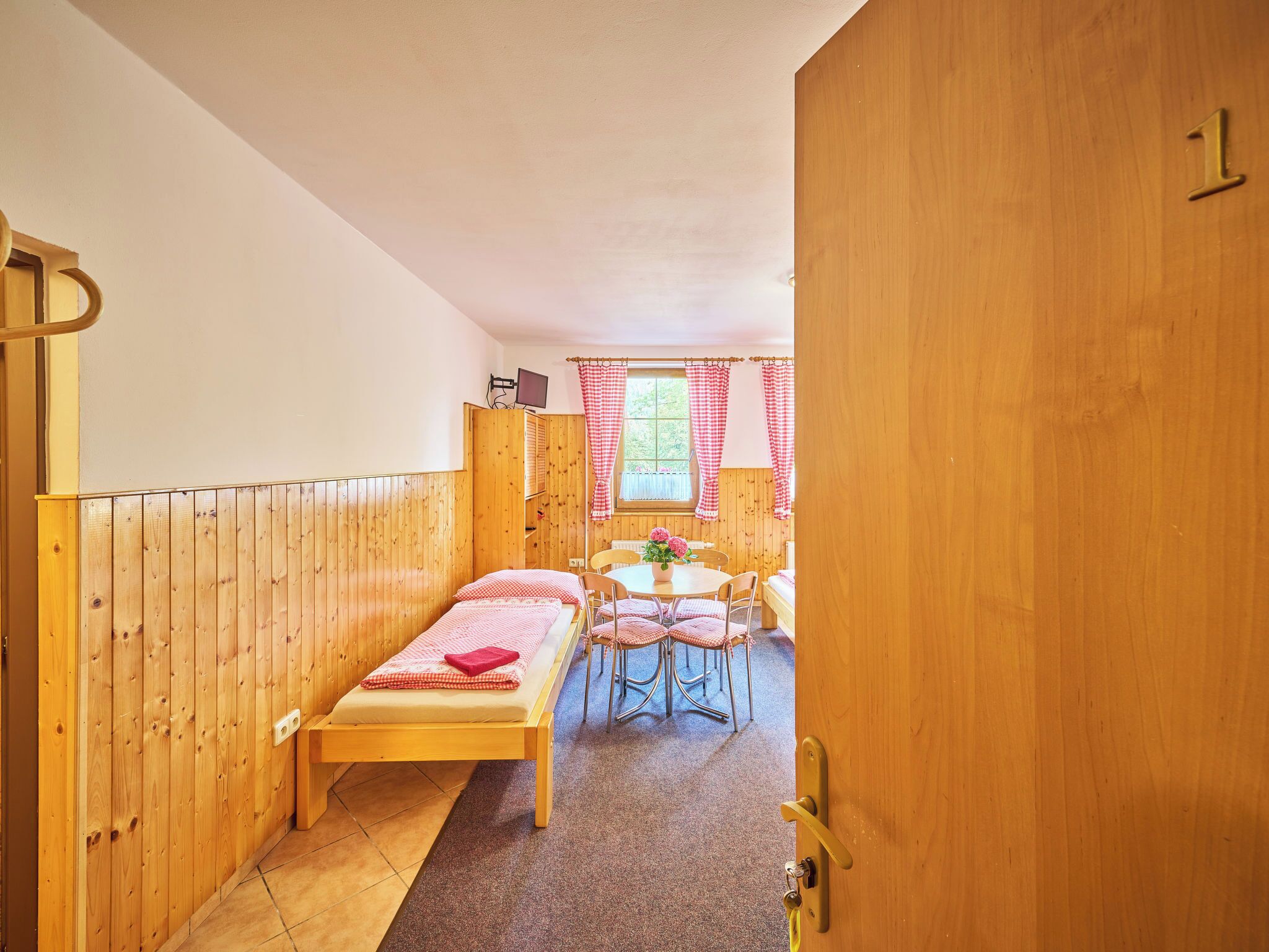 Una habitación de madera con una cama, una pequeña mesa con sillas y un ventanal con cortinas.
