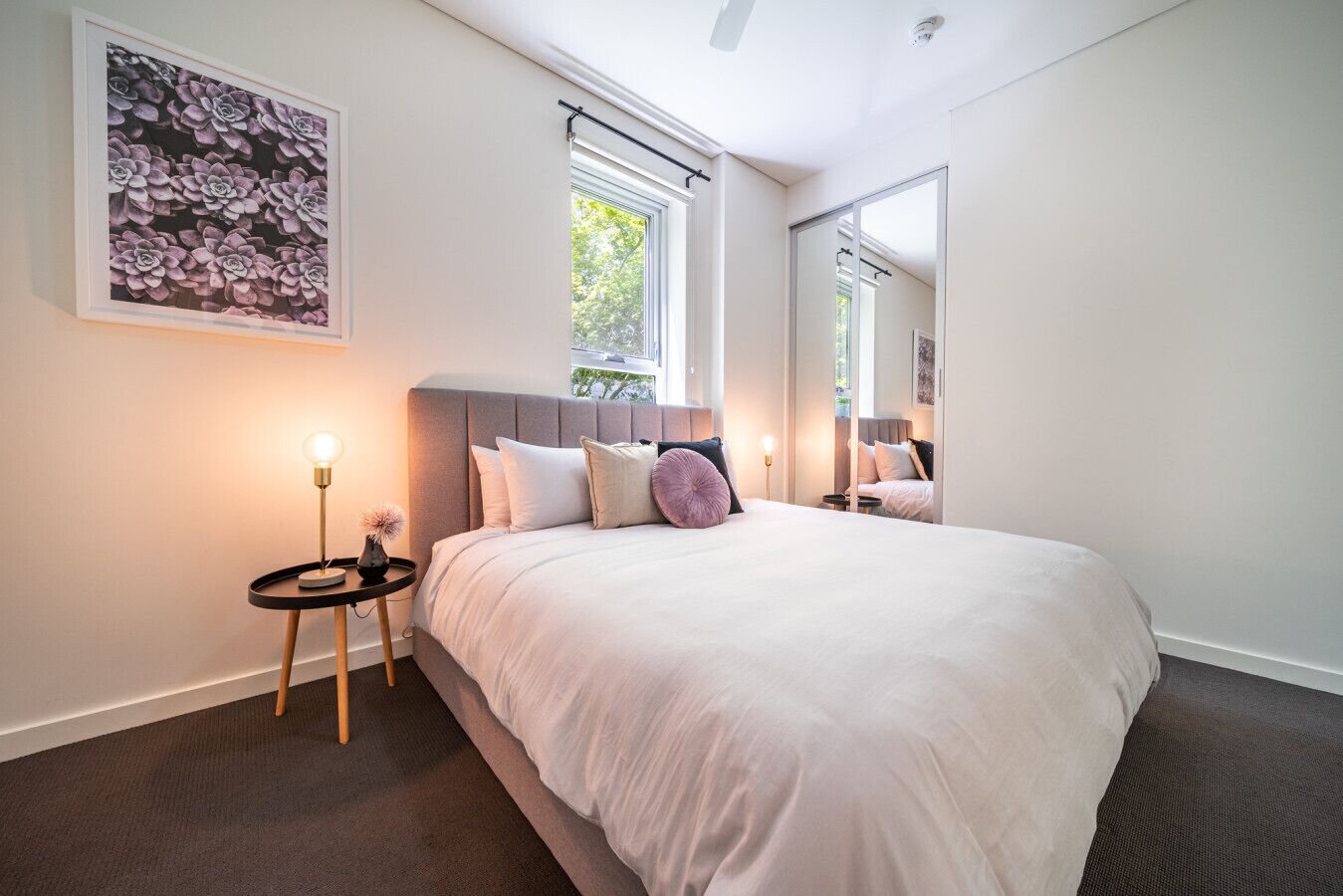 A modern bedroom with a large bed, a bedside table with a lamp, and a wall-mounted mirror.
