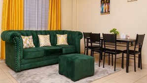 A green sofa with matching ottoman, a dining table with chairs, and a window with yellow curtains. - FURNISHED COZY ONE BEDROOM APARTMENT (Busia)