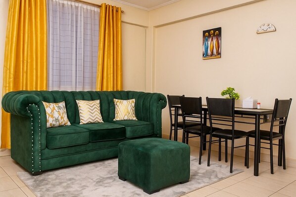 A green sofa with matching ottoman, a dining table with chairs, and a window with yellow curtains.