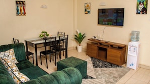 A living room with a green sofa, a dining table with chairs, a water dispenser, and a TV mounted on the wall. - FURNISHED COZY ONE BEDROOM APARTMENT (Busia)