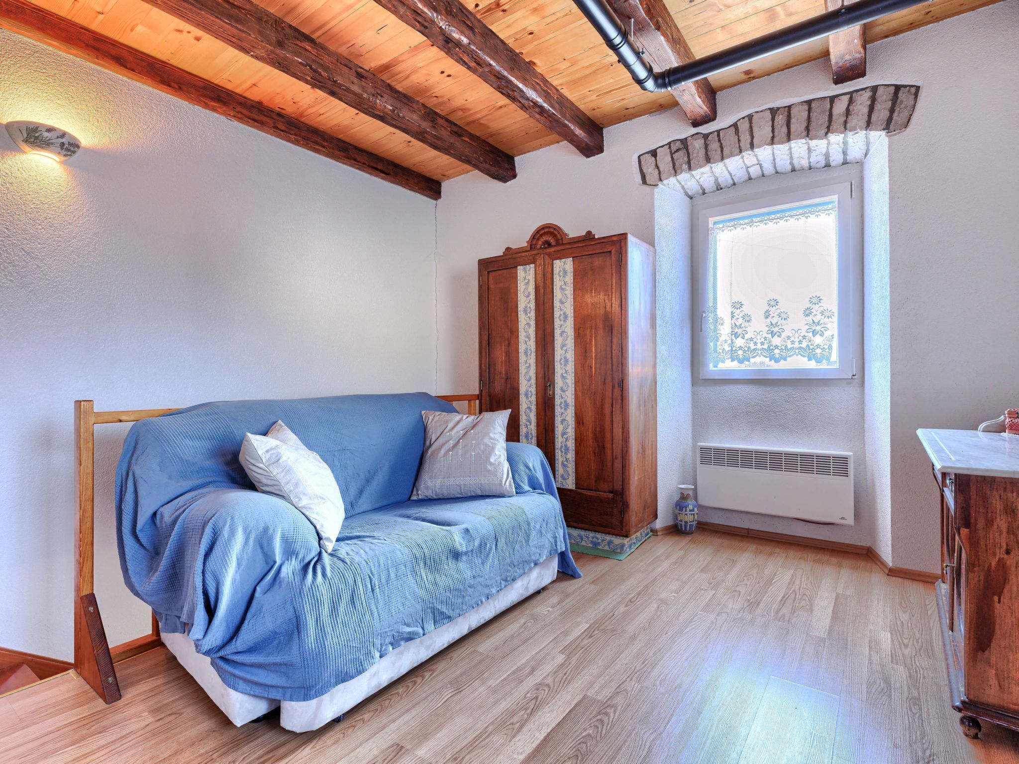 A cozy room with a wooden ceiling, a blue sofa, a wooden wardrobe, and a window with a decorative view.