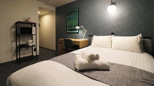 A hotel room with a neatly made bed, a desk with a lamp, a shelf with a mug, and a painting on the wall.