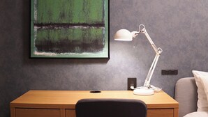 A room with a wooden desk, a white desk lamp, a grey textured wall, and a framed abstract painting.
