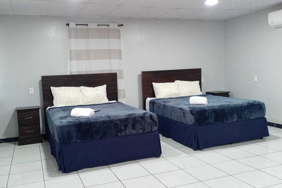 A hotel room with two beds, each with a dark blue bedspread and white pillows, a small bedside table, and a curtain.