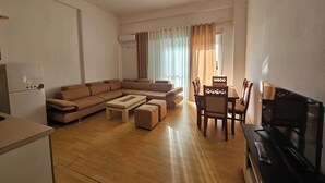 A modern living room with a sectional sofa, a dining table with chairs, a TV, and a kitchen area with a refrigerator. - Albania Dream Saranda Apartments (Saranda)
