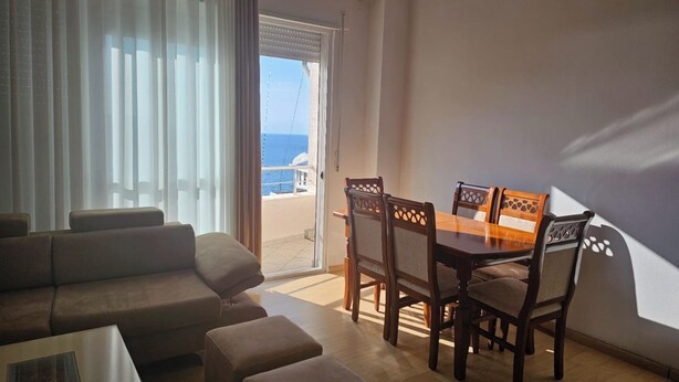 A living room with a sofa, ottomans, a dining table with chairs, and a view of the sea through a glass door. - Albania Dream Saranda Apartments (Saranda)