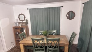 A dining area with a wooden table and four chairs, a small cabinet with decorative items, and a window with green curtains. - Spacious 4-bedroom apartment with WiFi, AC in charming Conway (Conway)