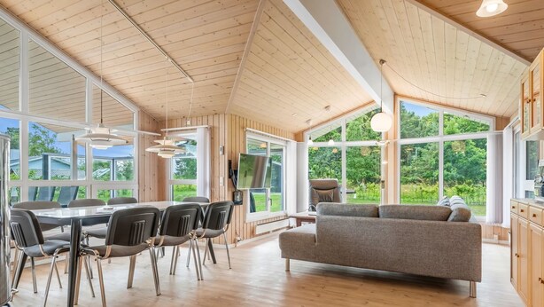 Une pièce spacieuse avec un plafond haut, des murs en bois, un coin repas avec une table et des chaises, un canapé et de grandes fenêtres donnant sur un paysage verdoyant.