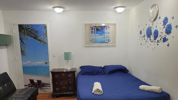 Une chambre à coucher comprenant un lit, une table de chevet, une commode, une horloge murale et une porte ornée d’une fresque représentant une plage.