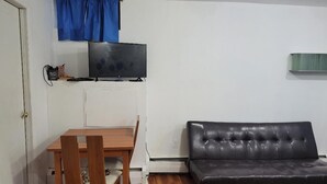 Un salon comprenant une table à manger en bois, deux chaises, un canapé en cuir noir, un téléviseur à écran plat et un rideau bleu.