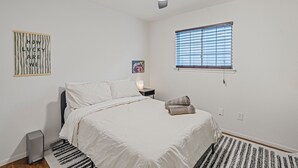 A bedroom with a bed, a ceiling fan, a window with blinds, a nightstand, a lamp, and a trash can. - Austin Escape w/ Pool, Hot Tub & Game Room – DT ,Bars, South Congress, Airport ! (Austin)