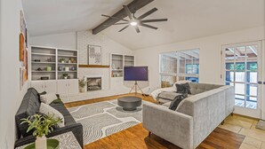A modern living room with a fireplace, built-in shelves, a grey sofa, and a large window with blinds. - Austin Escape w/ Pool, Hot Tub & Game Room – DT ,Bars, South Congress, Airport ! (Austin)