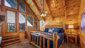A log cabin bedroom with a wooden bed, a nightstand, a lamp, and large windows. - NEW 6BR: Sleep 22| Pool| Views| Cinema| Hot Tub| A+ Location| Dog Friendly (Sevierville)