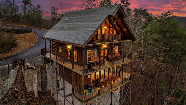 A multi-story log cabin with a balcony and outdoor seating area, surrounded by trees. - NEW 6BR: Sleep 22| Pool| Views| Cinema| Hot Tub| A+ Location| Dog Friendly (Sevierville)