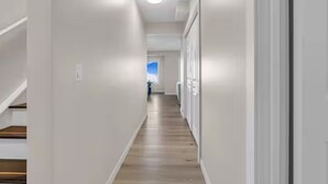 A narrow, well-lit hallway with wooden flooring and white walls, leading to a door at the end.