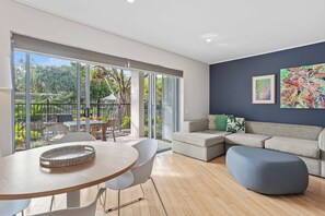 Een moderne woonkamer met een grote schuifpui die uitkomt op een terras, een grijze bank, een ronde tafel en een blauwe poef.