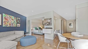 Un salon moderne avec un canapé d’angle, un pouf bleu, un coin repas comprenant une table en bois et des chaises blanches, et un téléviseur fixé au mur.