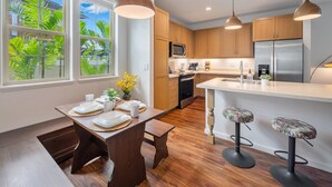 A modern kitchen with wooden cabinets, a stainless steel refrigerator, and a dining area with a wooden table and bar stools. - Ocean + Mountain Views|Beach 6 Min|Fire Pit|Gated (Waianae)