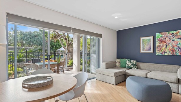 A modern living room with a large sliding glass door leading to a patio, a grey sofa, a round table, and a blue ottoman. - The Sand Resort Kingscliff - 1BR Plunge Pool - 7020 (Tweed)