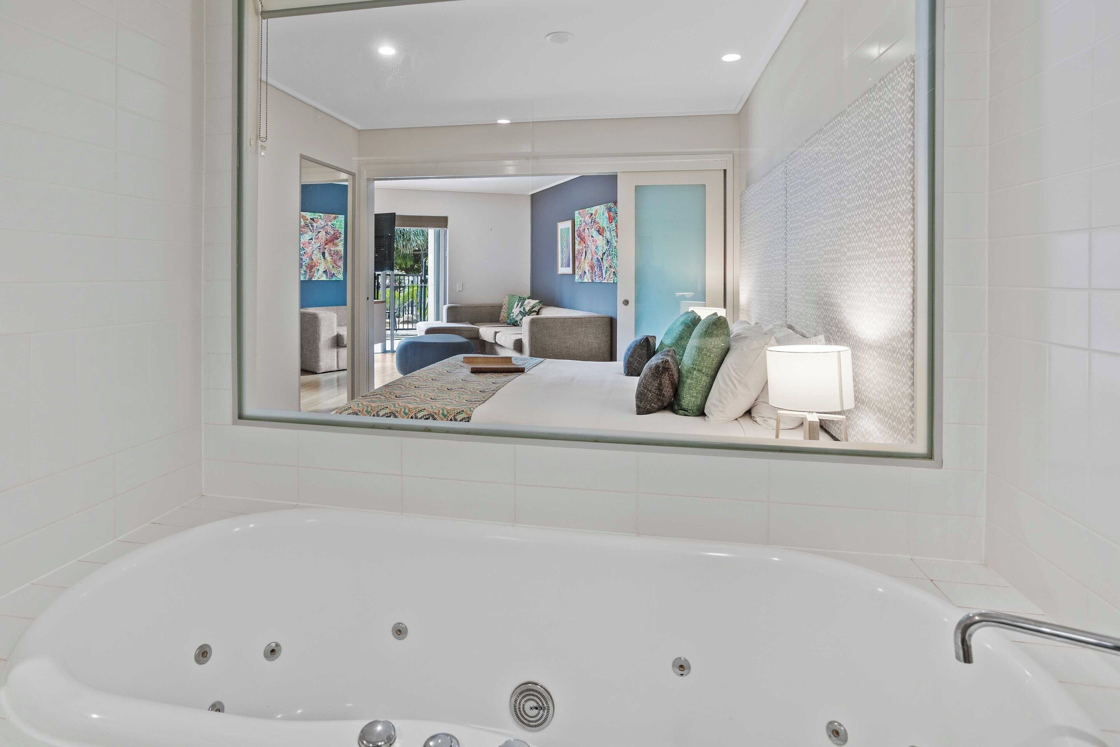 A modern bathroom with a large bathtub, a glass-enclosed shower area, and a visible bedroom with a bed and a nightstand.