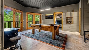 A pool table in a room with a wooden ceiling, a bar area, and a view of greenery through large doors.