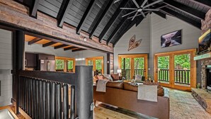A cozy cabin interior with a wooden ceiling, a fireplace, a large window, and a sofa.