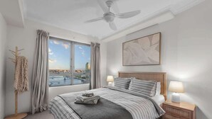 A hotel room with a bed, two bedside lamps, a ceiling fan, a window with curtains, and a city view.