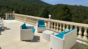 A terrace with white wicker furniture and blue cushions, overlooking a forested hillside.