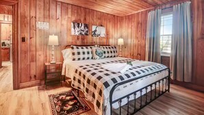 A wooden-paneled bedroom with a bed, two bedside lamps, a window with curtains, and a wall-mounted artwork. - The Whimsical Barn (Rocky Mount)