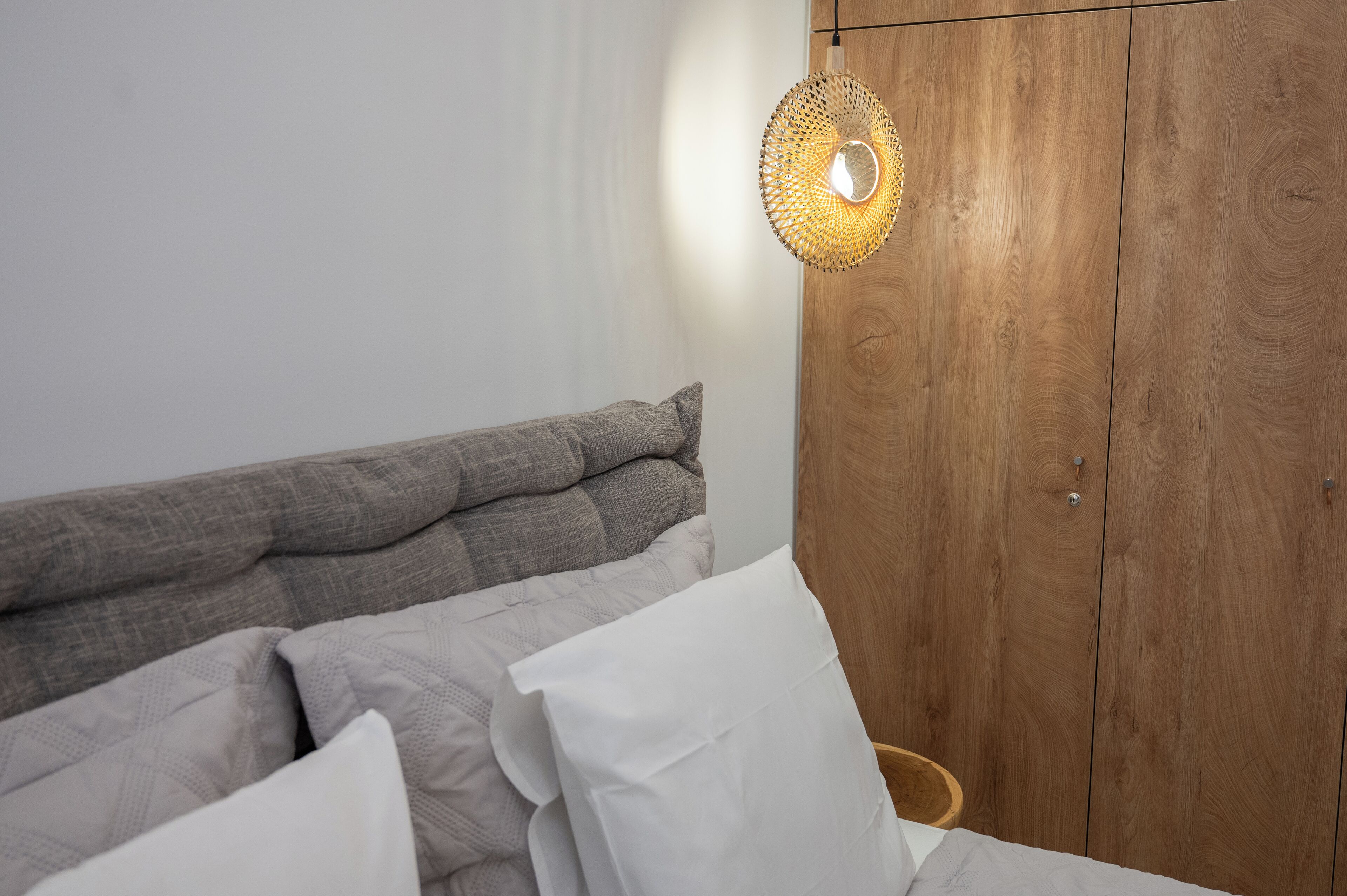 A bedroom with a wooden wardrobe, a bed with grey and white bedding, and a woven light fixture.