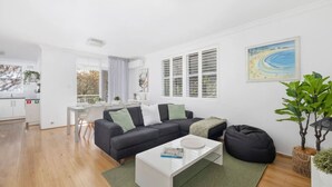 Een moderne woonkamer met een grijze bank, een witte salontafel en een zwarte relaxfauteuil. Er is een eethoek met een witte tafel en stoelen, en een keuken met witte kasten en een raam.