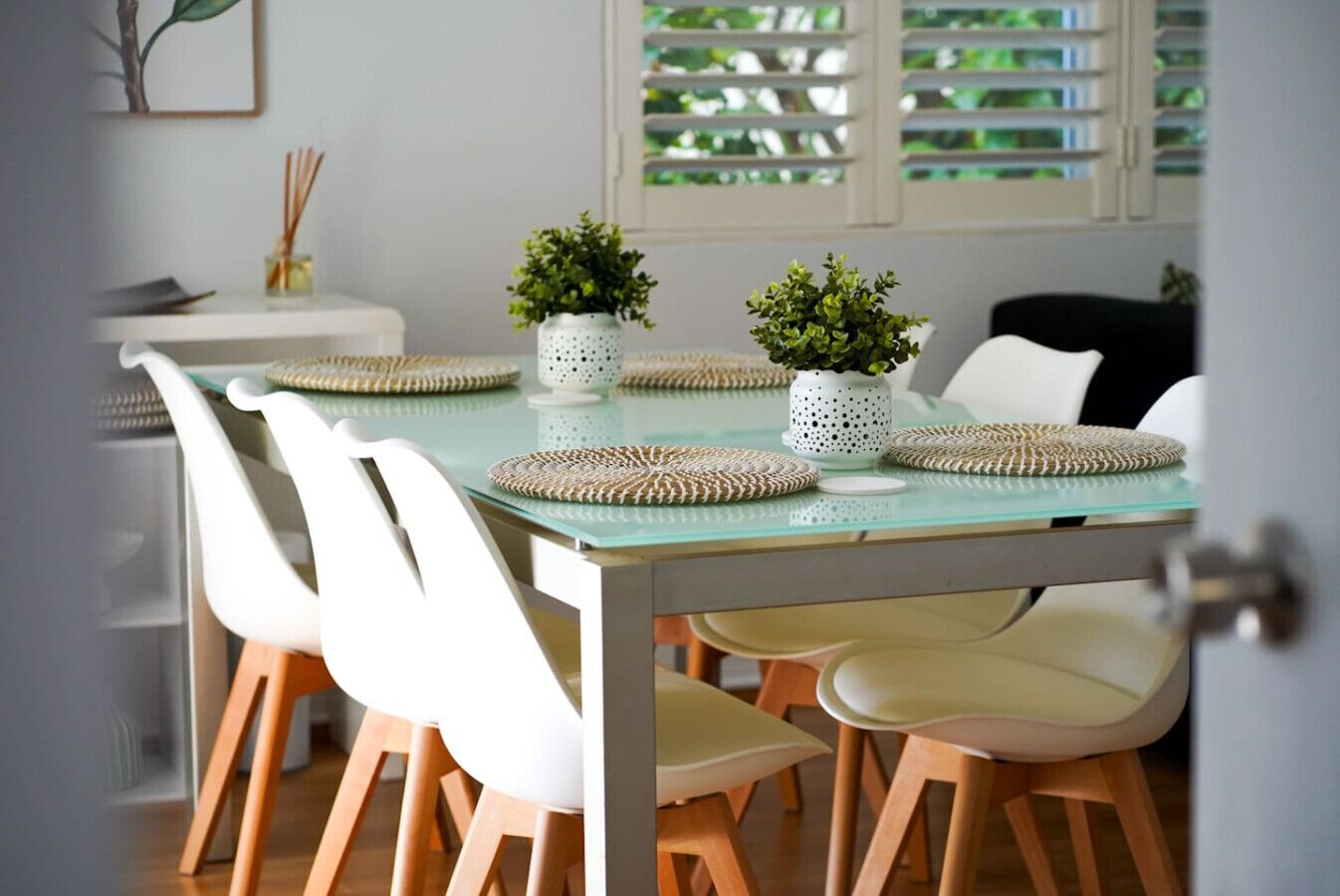 A dining area with a glass table, white chairs, woven placemats, and potted plants.