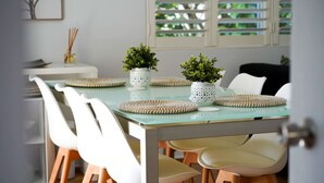 Een eethoek met een glazen tafel, witte stoelen, gevlochten placemats en potplanten.