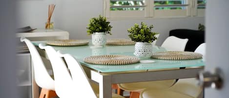 A dining area with a glass table, white chairs, woven placemats, and potted plants.