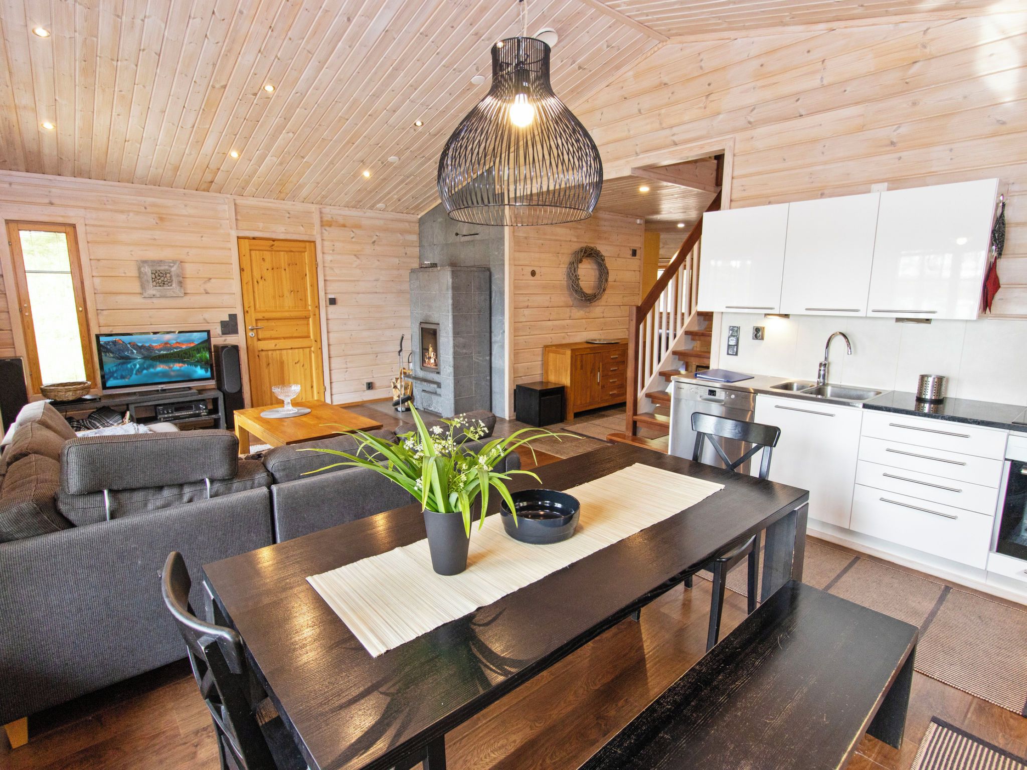 A modern interior with a wooden ceiling, a dining area with a black table, a sofa, and a kitchen with white cabinets.