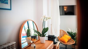 Une pièce avec un bureau en bois, un miroir, une orchidée en pot, une chaise en osier avec un coussin orange et un téléviseur fixé au mur.