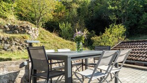 Une terrasse extérieure avec une table et quatre chaises, entourée d’un mur de pierre et d’une végétation luxuriante.