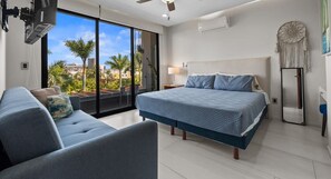 A modern hotel room with a large bed, a sofa, a ceiling fan, and a view of palm trees through large windows. - Quadrant 206: Pool, Gym & Padel – Walk to Beach! (Bucerías)