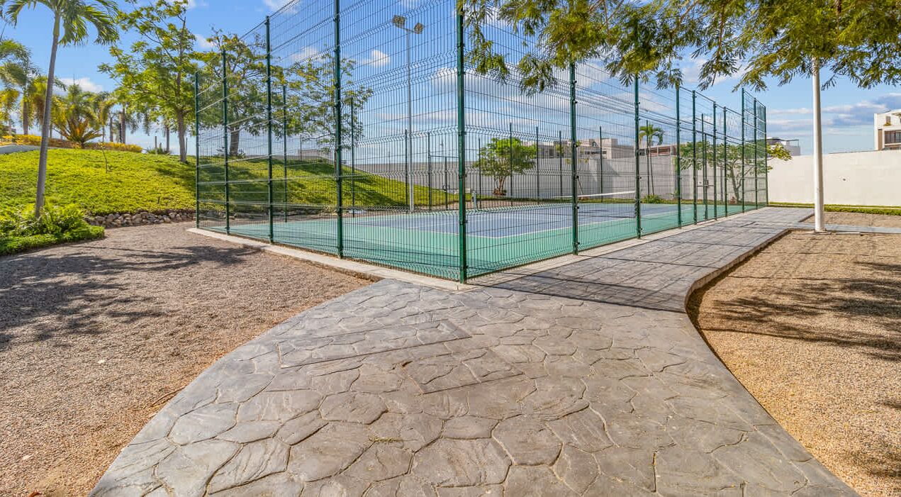 Ein Tennisplatz, umgeben von einem grünen Zaun, einer begrünten Anlage mit Bäumen und einem Kiesweg.