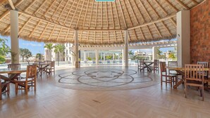 A spacious outdoor area with a thatched roof, wooden furniture, and a view of palm trees and buildings in the background.
