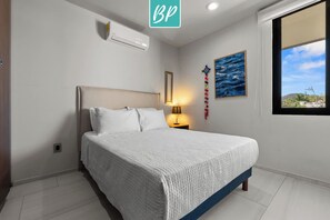 A bedroom with a bed, a bedside lamp, a wall-mounted air conditioner, a window with a view, and a framed picture on the wall. - Quadrant 206: Pool, Gym & Padel – Walk to Beach! (Bucerías)
