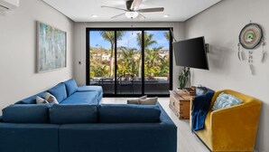 A living room with a blue sectional sofa, a yellow armchair, a flat-screen TV, and a large window with a view of palm trees and buildings.