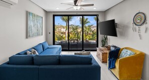 Una sala de estar con un sofá azul, un sillón amarillo, un televisor de pantalla plana y un ventanal con vistas a palmeras y edificios.