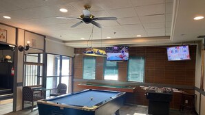 Una mesa de billar, una mesa de fútbol de mesa y un televisor mostrando un evento deportivo en una habitación con paredes de ladrillo y suelo de madera.