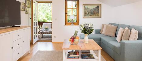 A living room with a TV, a coffee table with books and fruit, a sofa, and a window with a view of greenery.
