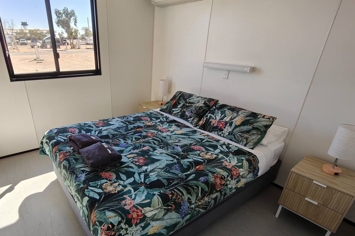 A bedroom with a bed, a nightstand, a window, and a view of the outside.